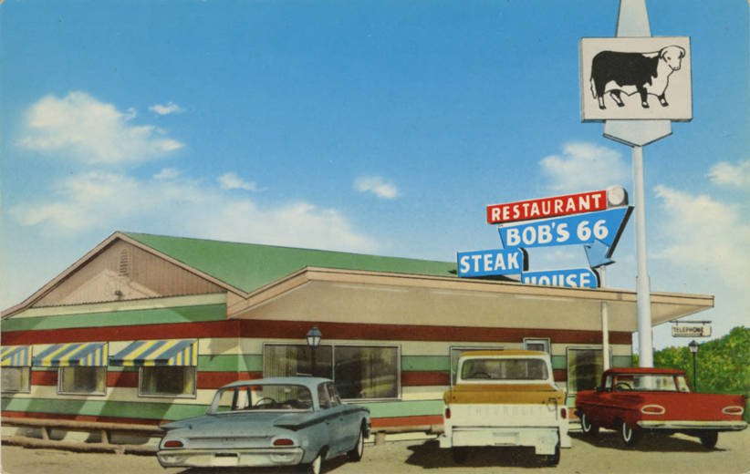 Vintage color postcard of Bob's 66 Steak House on old Route 66, with a striped awning, a steer sign on a tall pole and 1950s cars parked out front